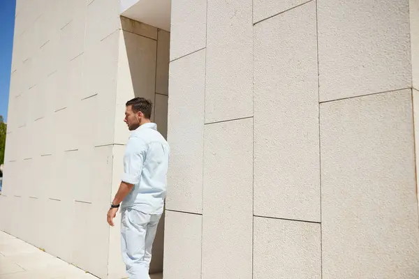 stock image Pensive young man in casual attire leaning against a minimalist, light-colored wall, reflecting in a bright, sunny outdoor setting.