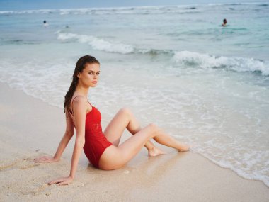 Kırmızı mayo giymiş bir kadın güneşten yanmış güzel bir vücutla kumların üzerinde oturur ve bir ada plajındaki tropiklerdeki okyanus kamerasına bakar. Yüksek kalite fotoğraf