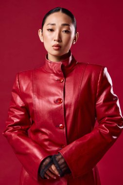 Kırmızı deri ceketli, siyah eldivenli, elleri kalçasında kırmızı arka planda poz veren Asyalı bir esmer. Şık görünümlü genç bir kadın..