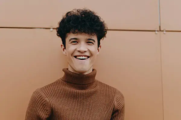 stock image Fashion portrait of a smiling young man in a brown turtleneck sweater, standing in front of an orange wall.