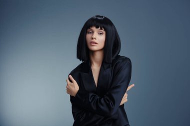 Siyah takım elbiseli güzel bir genç kadın profesyonel ışıklandırılmış stüdyo çekiminde gri bir arkaplana karşı poz veriyor, zarafet ve entelektüelliği vurguluyor.