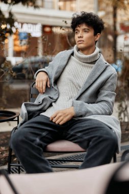 Süveter ve kot pantolonlu şık bir genç elinde bir fincan kahveyle bir masanın önünde oturuyor..