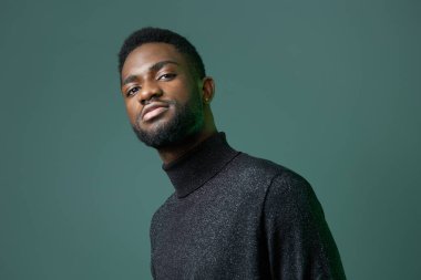 Siyah balıkçı yakalı kendinden emin genç adam kesinlikle canlı yeşil bir arka plana karşı duruyor, karizma ve tarz yayıyor..