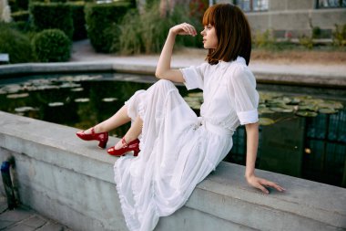 Zarif beyaz elbiseli, dingin kırmızı ayakkabılı, sakin bir göletin yanında oturan sakin bir kadın.