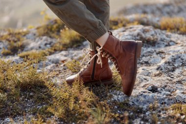 Kahverengi çizmeler kayalık tepede duruyor. Arka planda çimenler ve ağaçlar macera ve seyahat kavramını temsil ediyor.