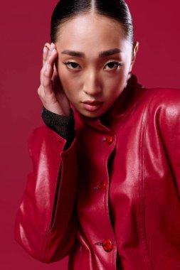Kırmızı deri ceketli Asyalı esmer, elleri başının üstünde kırmızı arka planda poz veriyor..
