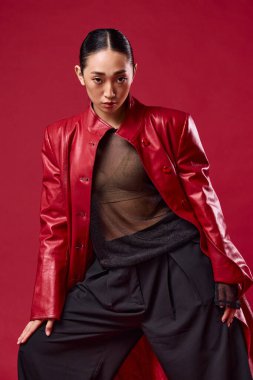 Kırmızı deri ceketli, siyah pantolonlu, kırmızı arka planda poz veren Asyalı kadın. Elleri kalçasında duruyor, kendine güveni ve tarzı var..