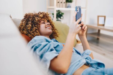 Cep telefonu tutan mutlu kadın, evdeki kanepede dinleniyor.