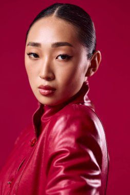 Kırmızı deri ceketli Asyalı esmer, kırmızı arka planda poz veriyor, elleri kalçasında ve gözleri yana bakıyor..