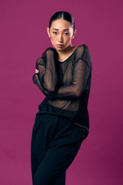 Mor arka planda poz veren siyah elbiseli Asyalı manken elleri kalçasında ve kafası yana yatık..