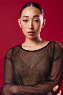 Saf siyah bluzlu Asyalı kadın kırmızı arka planda poz veriyor. Kolları çapraz ve elleri kalçasında. Model.