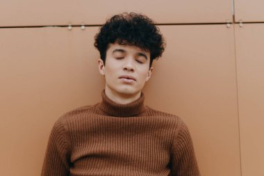 Kıvırcık saçlı, kahverengi bir kazak giyen, gözleri kapalı ve elleri ceplerinde bir duvara yaslanmış şık bir genç..