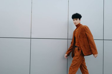 Kahverengi takım elbise ve balıkçı yaka giyen şık bir genç adam modern gri bir duvar boyunca yürüyor, kentsel moda ve entelektüelliği somutlaştırıyor.