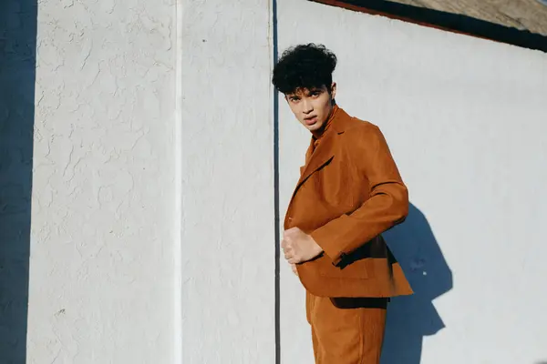 stock image A young man dressed in a brown suit poses confidently against a textured white wall The sunlight highlights his features and creates dramatic shadows