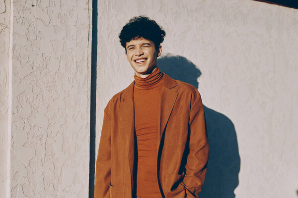 A cheerful young man stands confidently in front of a pale wall, dressed in a brown ensemble The warm sunlight highlights his joyful expression and creates a playful shadow