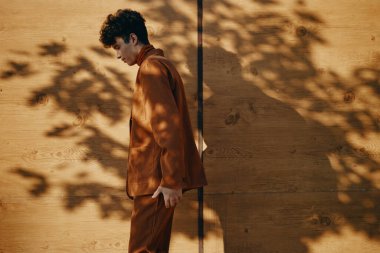 Kahverengi takım elbiseli şık bir genç, yaprakların yumuşak gölgeleriyle çevrili ahşap bir duvara yaslanır.