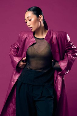 Mor ceketli, siyah pantolonlu, elleri kalçasında, göğsünde mor arka planda poz veren Asyalı bir kadın..