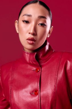 Kırmızı deri ceketli ve rujlu genç Asyalı kadın elleri kalçasında kırmızı arka planda poz veriyor. Moda esmer modeli..