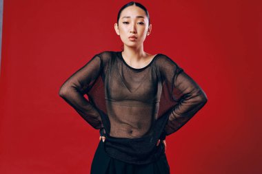 Saf üst ve siyah pantolonlu Asyalı manken elleri kalçalarında kırmızı arka planda poz veriyor..