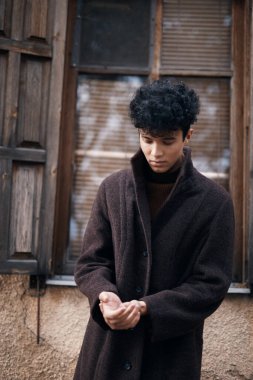 Kahverengi paltolu şık bir genç, arka planda ahşap kapı ve pencereyle eski binanın önünde duruyor..