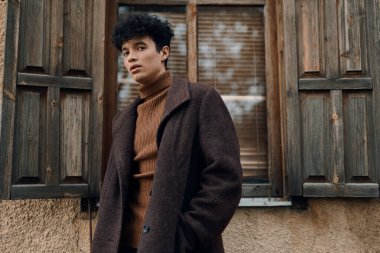 Kahverengi ceketli ve balıkçı yaka kazaklı moda adam pencerenin önünde duruyor..