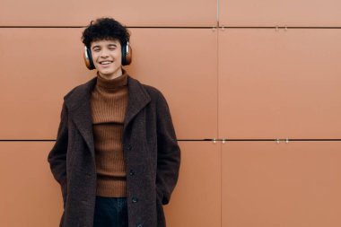 Turuncu duvara karşı duran kulaklıklı bir gencin moda portresi. Kafasında kahverengi bir ceket, eşarp ve bere var..