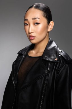Moda Asyalı kadın siyah deri ceketli stüdyo resmi gri arka plan. Zarif bir Asyalı kadın, bir stüdyo fotoshoot için şık bir siyah deri ceket içinde, nötr gri bir renge karşı duruyor.