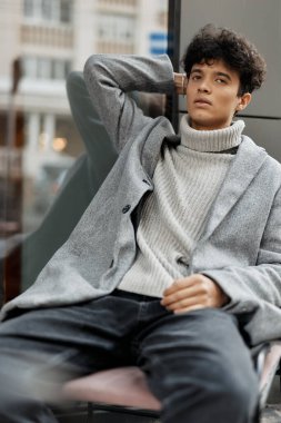 Gri bir kazak ve siyah balıkçı yaka giyen genç bir adamın moda portresi. Elleri başının üzerinde, pencerenin önünde bir sandalyede oturuyor..