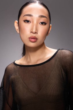 Gri bir arkaplan önünde poz veren katıksız bluzlu Asyalı bir kadın..