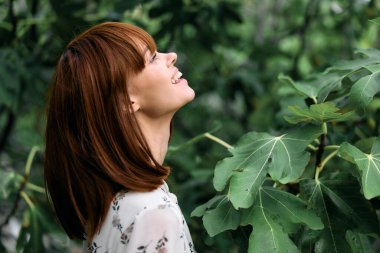 Büyüleyici kızıl saçlı kadın dingin ormandaki yemyeşil ağaç gölgesine bakıyor.