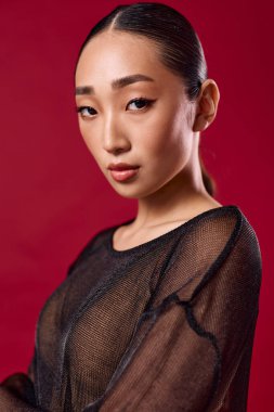 Saf siyah bluzlu Asyalı kadın kırmızı arka planda poz veriyor gözleri kapalı ve elleri kalçasında.