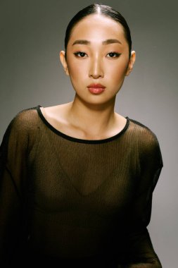 Gri bir arkaplan önünde poz veren katıksız bluzlu Asyalı bir kadın..