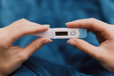 Kolunda mavi bir battaniye ve arka planında mavi bir yastık olan elektronik bir termometre tutan bir kişi..