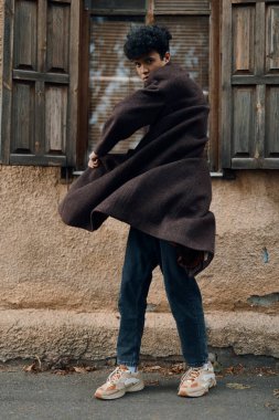 Moda, kahverengi paltolu şık bir adam bir caddenin kenarında duruyor elleri ceplerinde ve başı yana yatık..