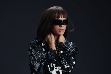 A woman in sunglasses and a sequinned dress, with her hands on her face and head tilted to the side, poses against a black background, exuding glamour and sophistication clipart