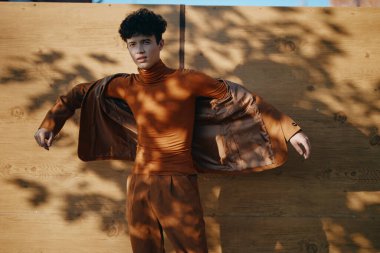 Kahverengi takım elbiseli şık bir erkek model ahşap bir arka plana karşı duruyor, özgüven ve yaratıcılık sergiliyor.