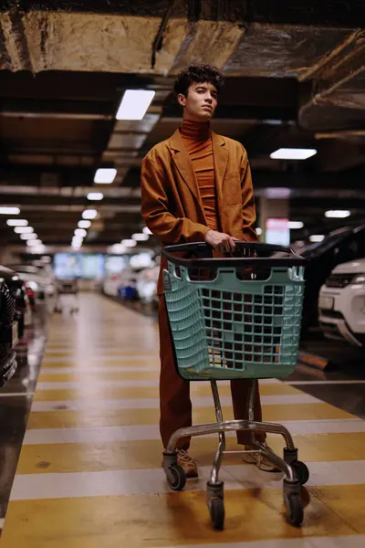 stock image A stylish young man in a brown suit stands with a shopping cart in a dimly lit parking garage The environment includes parked cars and vibrant yellow lines