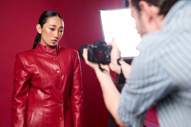 Kırmızı deri ceketli Asyalı esmer kadın fotoğraf çeken bir adamla fotoğraf çektirmek için poz veriyor. kırmızı arkaplan ve ışık resme dramatik bir etki katar.