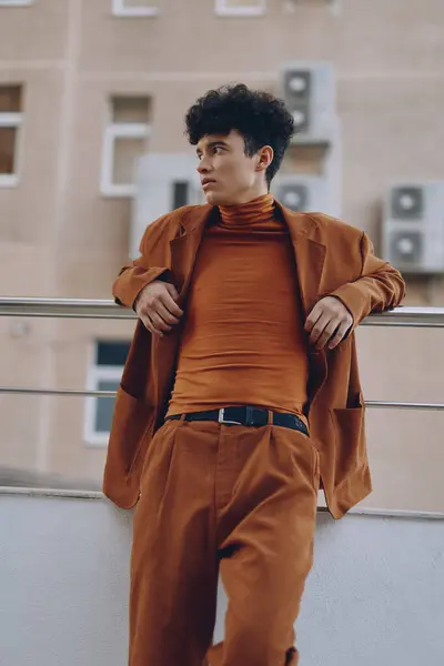 stock image A stylish young man dressed in an orange turtleneck and matching blazer, leaning against a railing with a neutral wall in the background