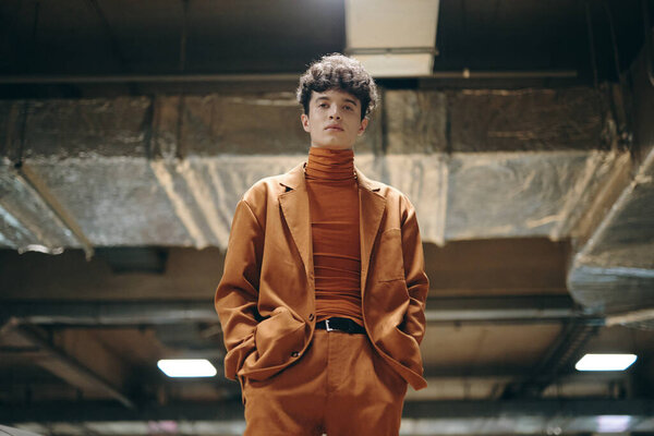 A fashionable male model wearing an orange suit and turtleneck poses in an urban, industrial environment with a contemporary vibe