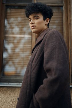 Kıvırcık saçlı, kahverengi ceketli, pencerenin önünde duran ve yüzünün kenarına bakan genç bir adamın moda portresi..
