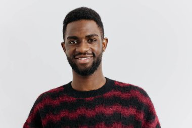 Kırmızı ve siyah kazaklı kendinden emin genç bir adam tarafsız bir arkaplanda duruyor, kararlı bir şekilde ileriye bakıyor..