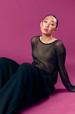 Saf bluzlu, siyah pantolonlu, mor arka planda oturan, gözleri kapalı ve elleri kalçasında olan Asyalı bir kadın..