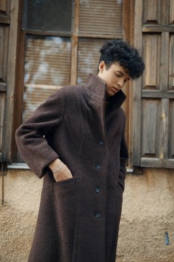 Kahverengi paltolu şık bir genç adam klasik bir binanın önünde elleri kalçasında ve başı yana yatmış şekilde poz veriyor..