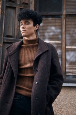 Kahverengi ceketli ve balıkçı yaka kazaklı şık bir genç arka planda penceresi olan eski bir binanın önünde dikiliyor..