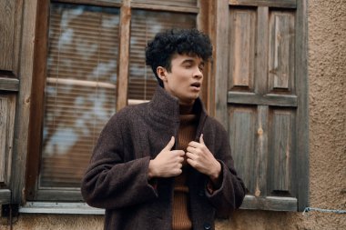 Eski binanın önünde duran kıvırcık saçlı genç adam. Açık pencereli, kahverengi ceketli..