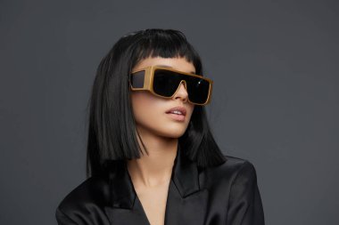 Siyah saçlı, güneş gözlüklü, siyah ceketli, altın logolu şık bir kadın koyu renk saçlı, güneş gözlüklü, siyah ceketli, önünde altın bir amblem olan bir kadın.