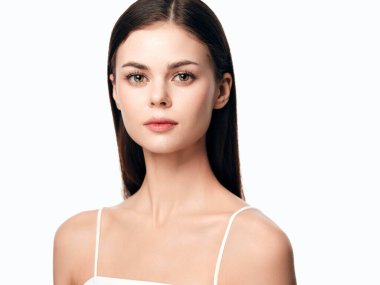 Uzun kahverengi saçlı, beyaz kolsuz bluz giyen, beyaz arka planda poz veren çekici bir genç kadın..