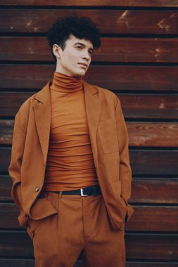 Pas rengi bir takım elbise ve balıkçı yaka giyen genç bir adam ahşap bir duvara karşı duruyor. Kendine güveni ve modası geçmiş moda estetiğini sergiliyor.