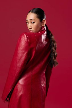 Kırmızı deri ceketli Asyalı şık esmer kadın elleri kalçasında kırmızı arka planda poz veriyor. Saçını at kuyruğu yapmış..